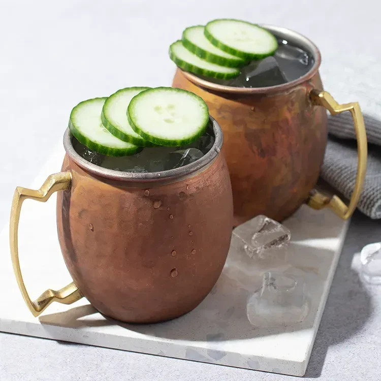 Two hammered copper Moscow Mule mugs with fresh cucumber and ice.