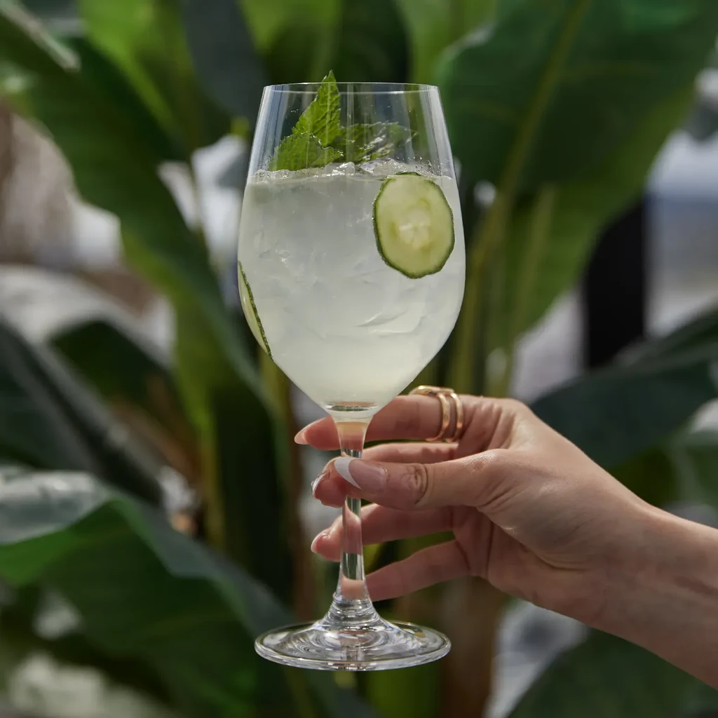Hand holding iced drink with mint, cucumber, against green plants.