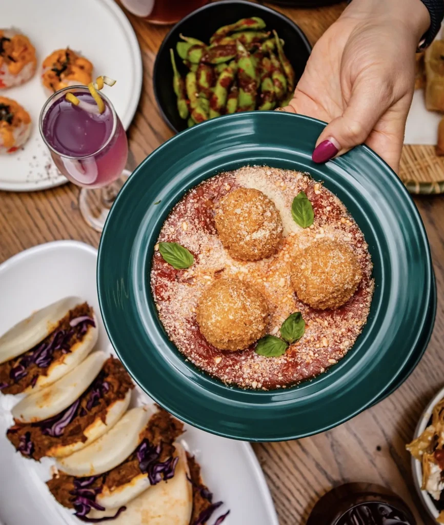 Hand holds bowl of breaded spheres, sauce, cheese; various dishes on table.