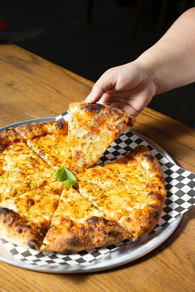 Hand lifting a cheesy pizza slice from a checkered tray with basil.