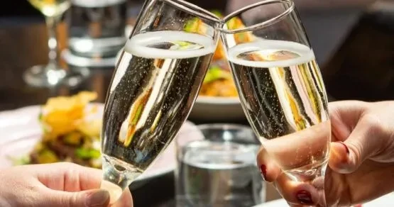 Two hands clink sparkling champagne flutes in a blurred dining setting.