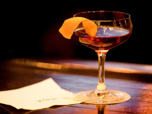 Deep reddish-brown cocktail with vibrant orange peel in coupe glass.