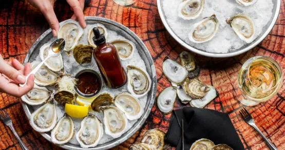 Hands spooning sauce onto fresh oysters on ice with wine.