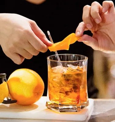 Hands garnishing an amber cocktail with an orange twist.