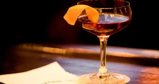 Deep reddish-brown cocktail in coupe glass with twisted orange peel.