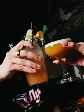 Hands with cocktails toast in a moody bar with neon signs.