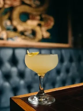 Pale gold cocktail with lemon twist in coupe glass on bar.