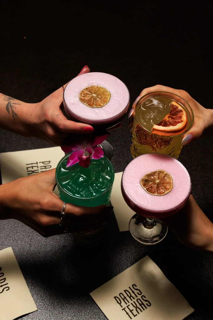 Four hands toast distinct cocktails; 'PARIS TEXAS' napkins visible.