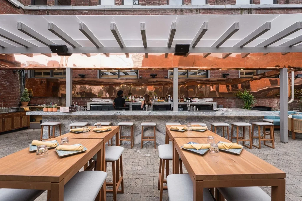 Outdoor bar with copper backsplash, long wooden dining tables, and staff.