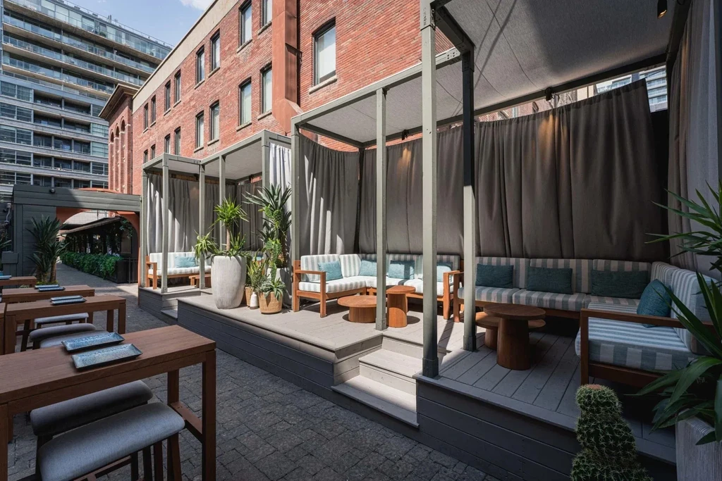 Outdoor restaurant patio with cabana seating, high tables, and city buildings.