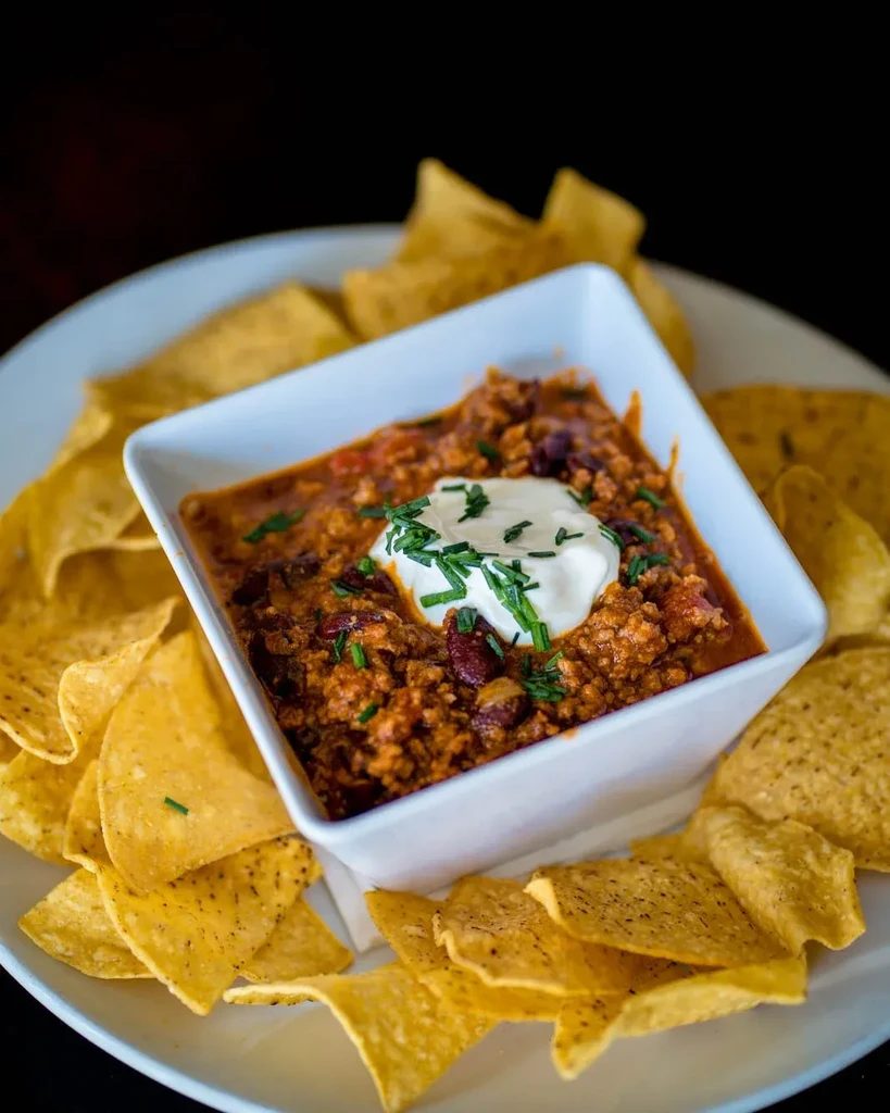 Hearty chili in a square bowl with sour cream, chives, and chips.