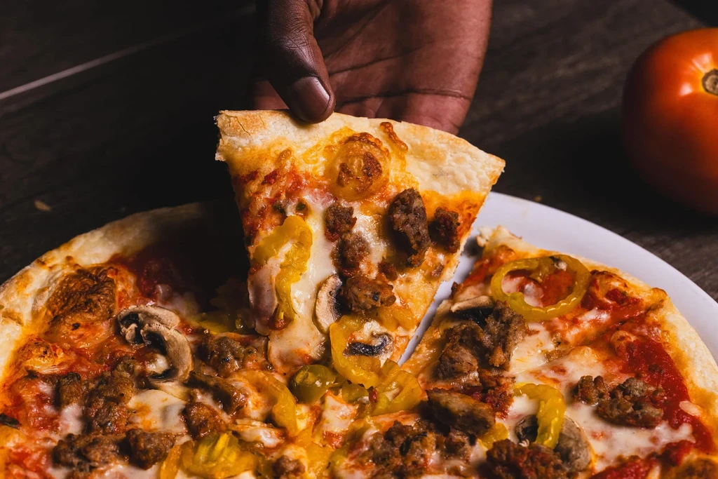 Hand lifting a pizza slice topped with meat, cheese, mushrooms, peppers.