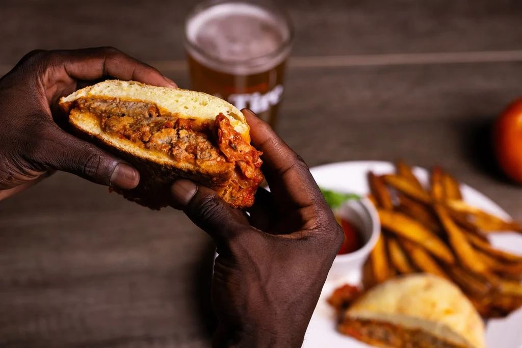 Dark-skinned hands hold a half-eaten red-sauce meat sandwich, fries and beer.