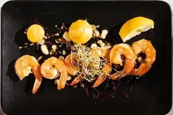 Five cooked shrimp, microgreens, yellow sauce, and lemon on black plate.