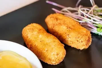 Two golden-brown fried appetizers, sauce, and garnish on dark plate.