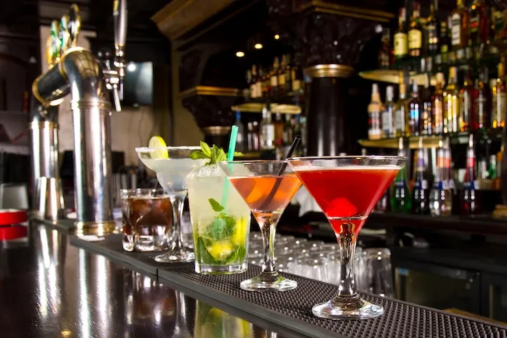 Five colorful cocktails on a dark bar counter with bottles behind.