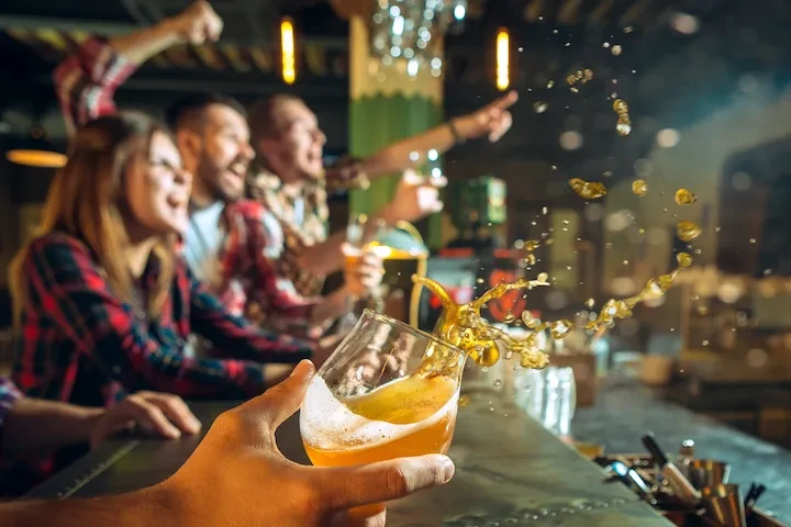 Hand holds beer splashing from glass, a cheering crowd blurs in background.