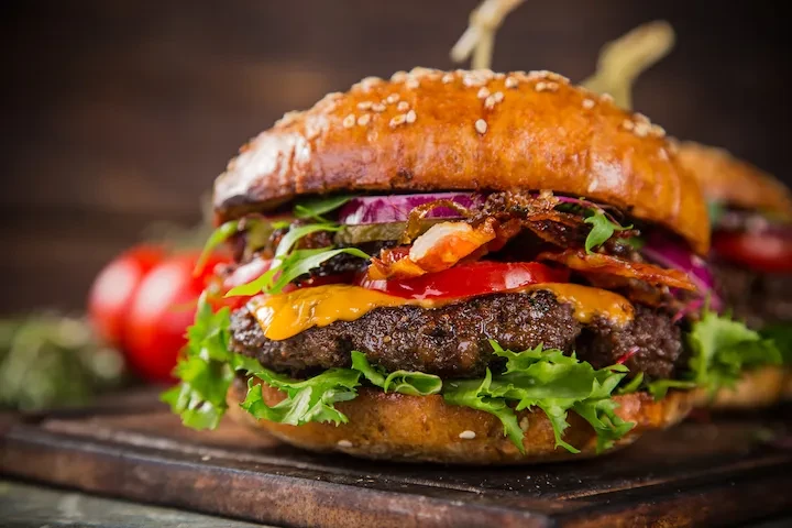 Gourmet beef burger with bacon, cheese, and vegetables on a wooden board.