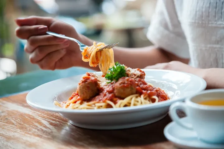 Hand lifting spaghetti and meatballs garnished with cheese and parsley.