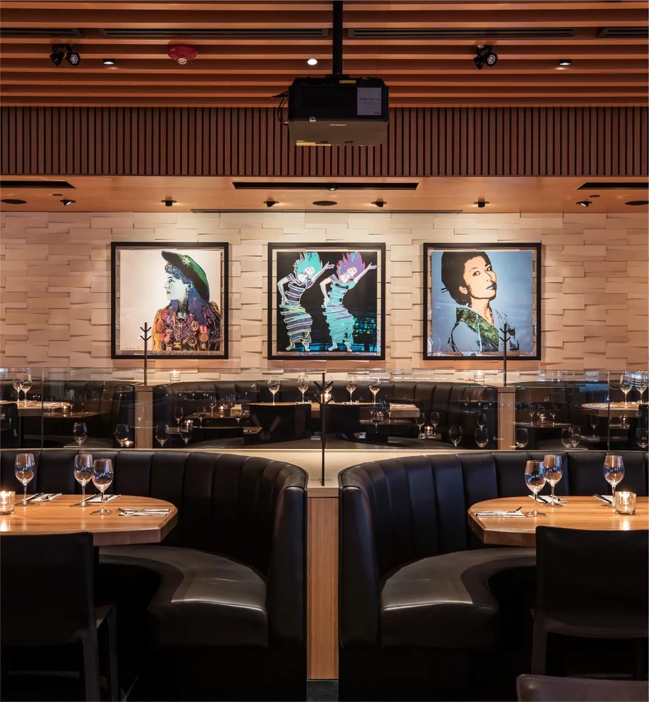 Sophisticated restaurant with black booths, set tables, pop art, and wooden ceiling.