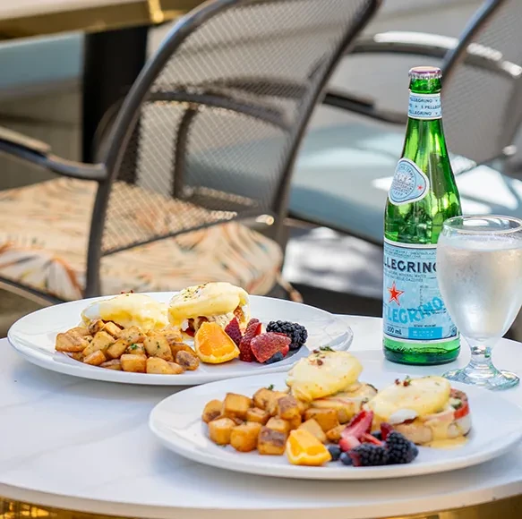 Two plates of Eggs Benedict, roasted potatoes, fruit, and drinks outdoors.
