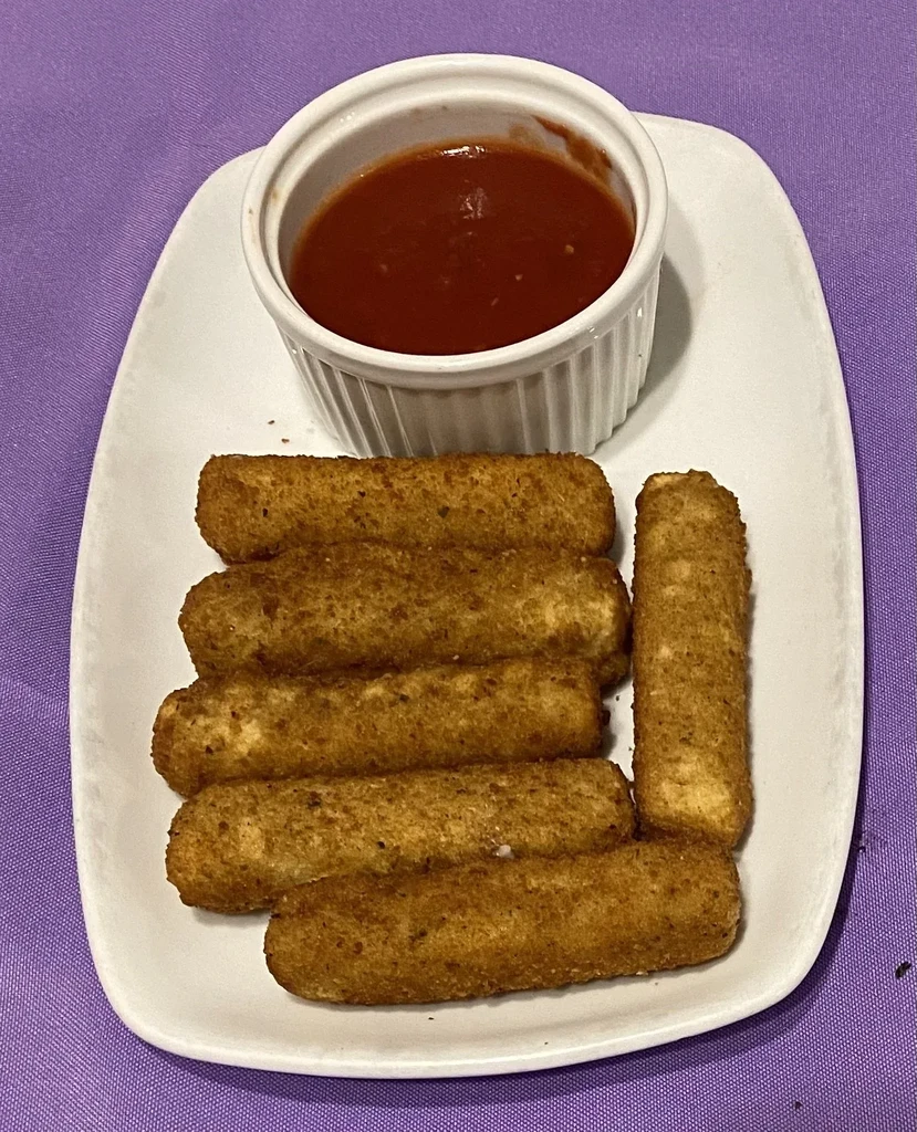Six crispy mozzarella sticks and red sauce on a plate, purple background.