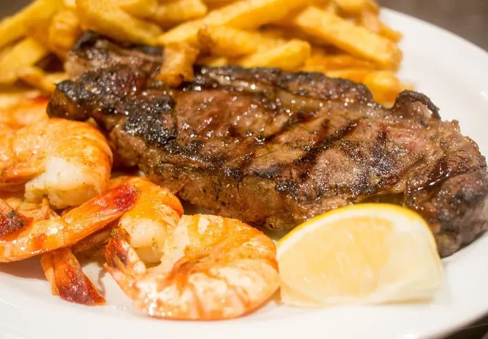 Grilled steak with shrimp, lemon, and french fries on a white plate.