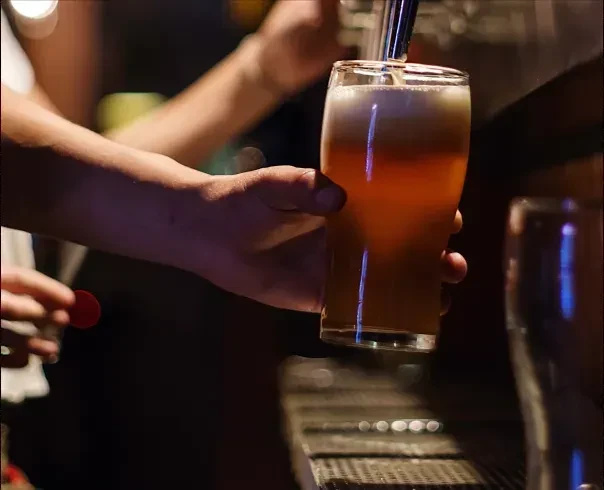 Hand holds glass of frothy golden beer being poured from a tap.