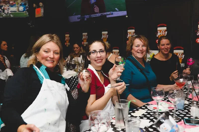 Four women in aprons paint wine glasses at a lively bar.