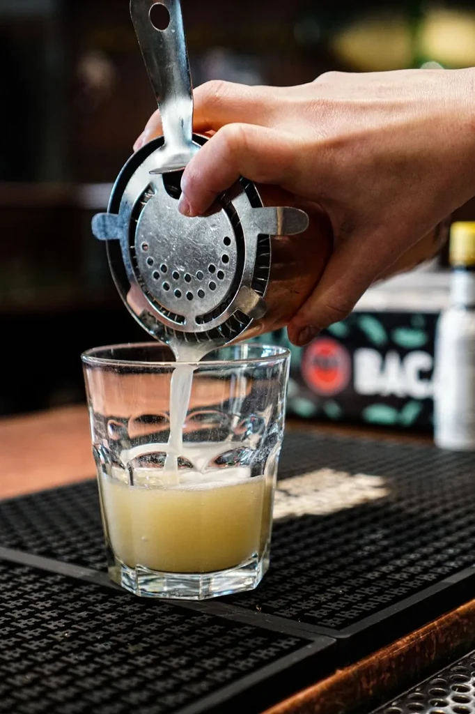 Close-up of hand straining frothy yellow cocktail into glass.