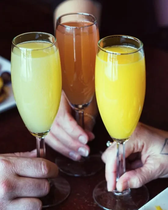Three hands raise champagne flutes filled with mimosas for a toast.