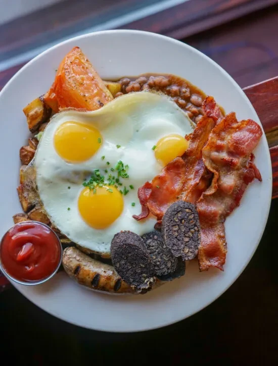 Plate of fried eggs, crispy bacon, sausage, beans, tomato, and black pudding.