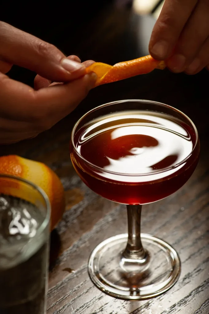 Dark red cocktail in coupe glass with hands expressing orange peel.