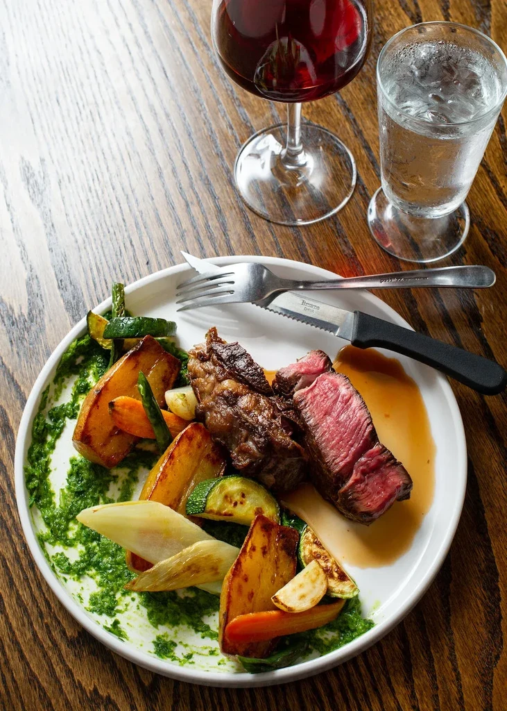 Medium-rare steak, roasted vegetables, red wine, and water on a wooden table.