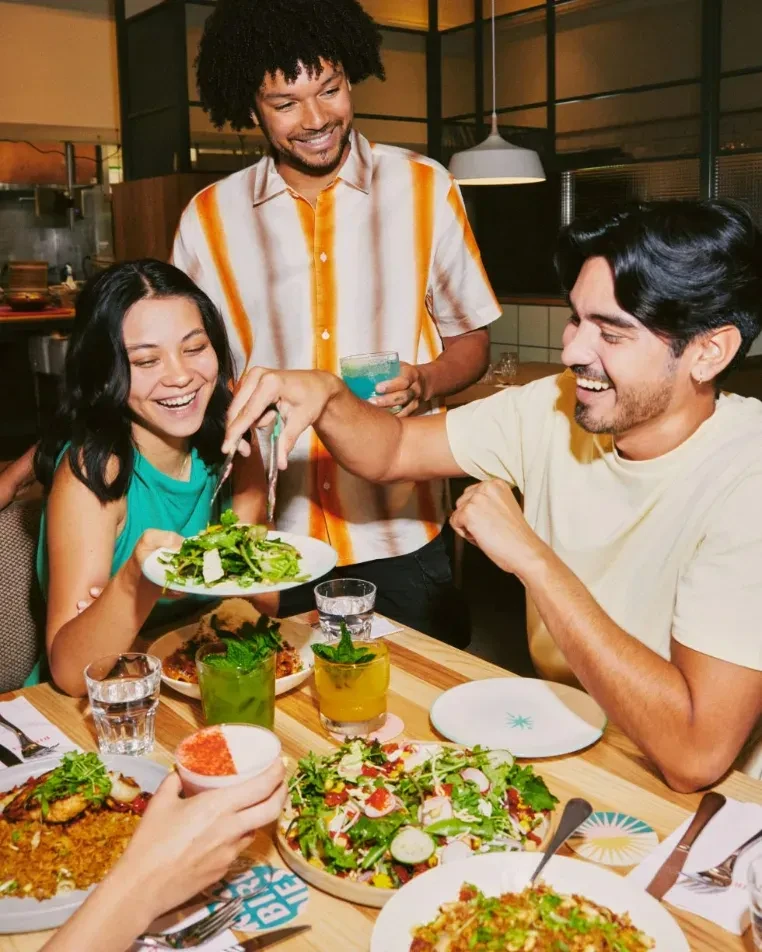 Three young adults laughing and sharing a meal at a restaurant.