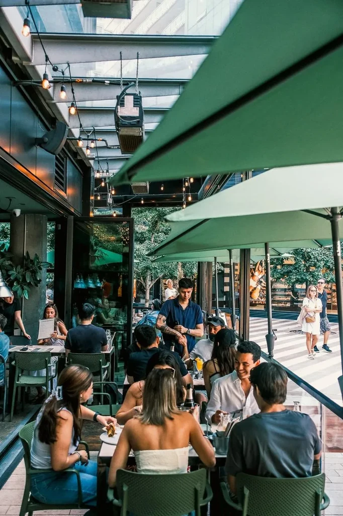 Busy restaurant patio with diners, umbrellas, string lights, and a waiter.