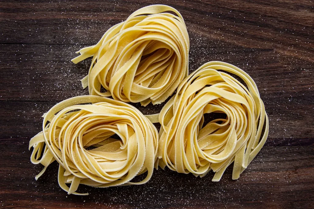 Three nests of raw yellow pasta on dark wood, sprinkled with flour.