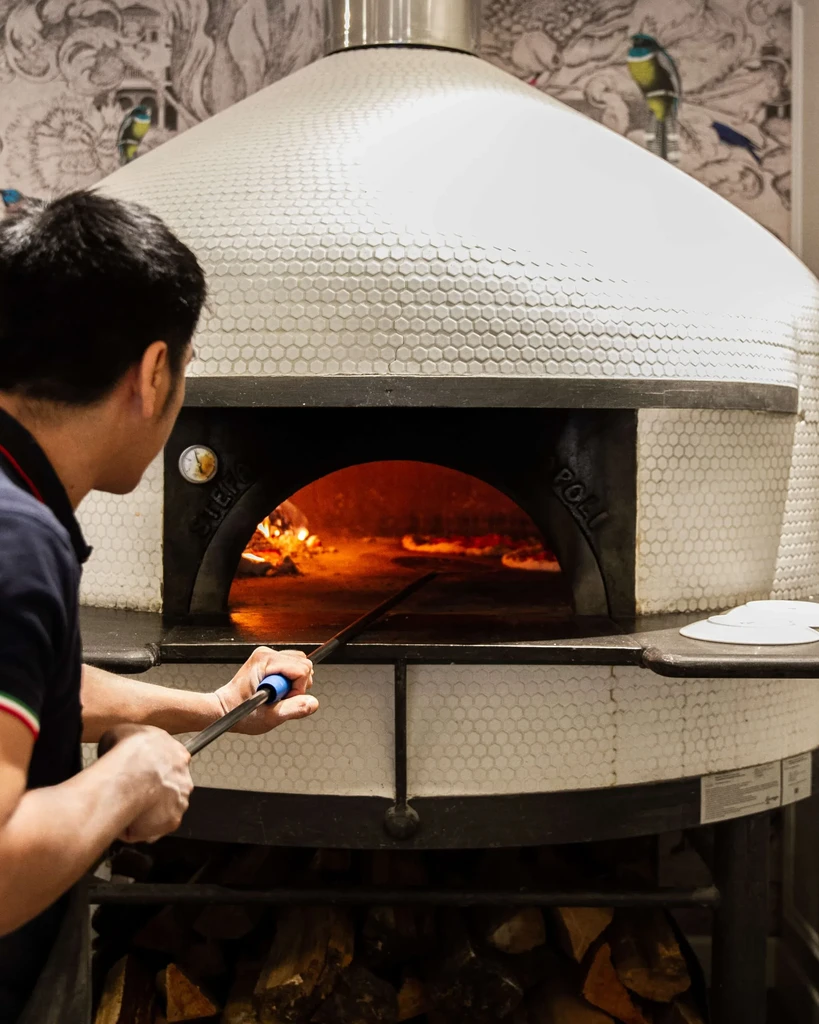 Chef baking pizzas with a peel in a tiled wood-fired oven.