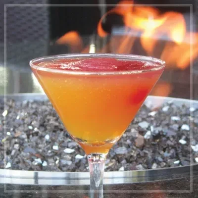 Effervescent orange-red cocktail with cherry, blurred fire flames in background.