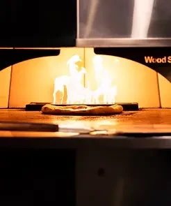 Pizza cooking inside a glowing wood-fired oven with bright flames.