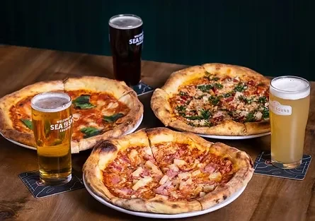 Three freshly baked pizzas and three beers on a wooden table.