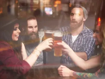 Four smiling people toast with beer glasses in a dim bar.