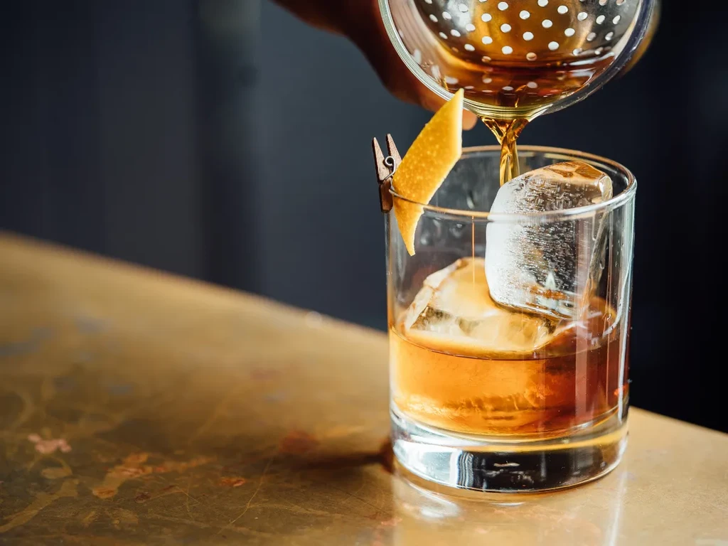 Hand pouring golden cocktail with ice and citrus peel garnish.