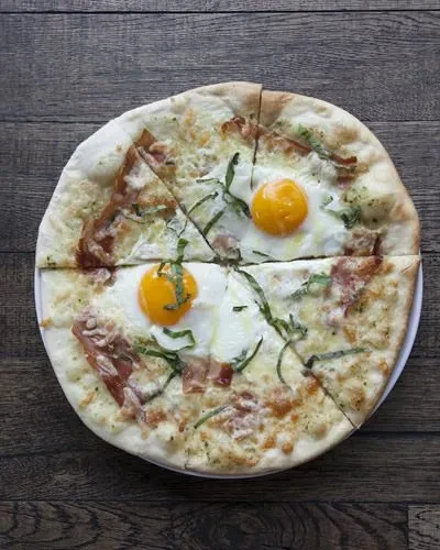 Top-down pizza with white sauce, ham, herbs, and two sunny-side-up eggs.