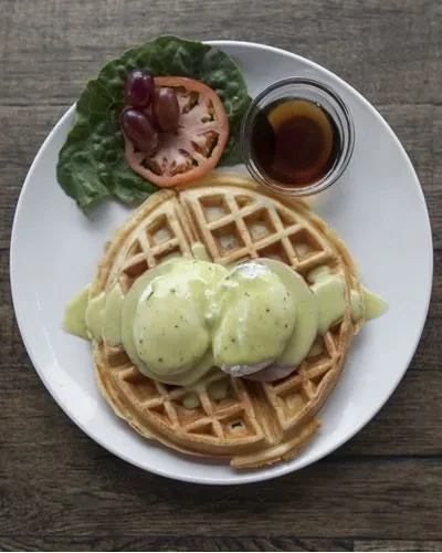 Golden-brown waffle with poached eggs, green hollandaise, ham, and side salad.