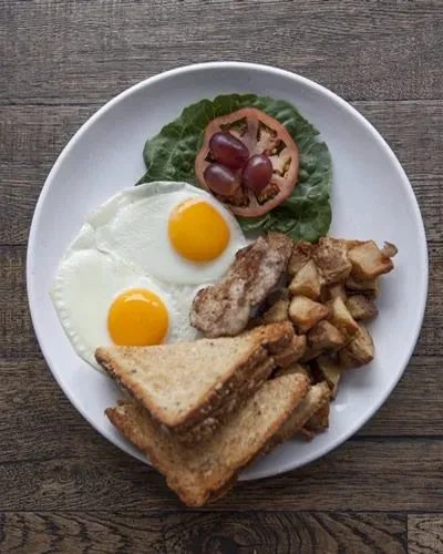 Breakfast plate with fried eggs, toast, home fries, meat, tomato, grapes.