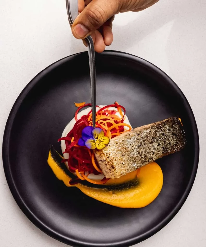 Chef's hand plating seared fish with vibrant vegetables and sauce.