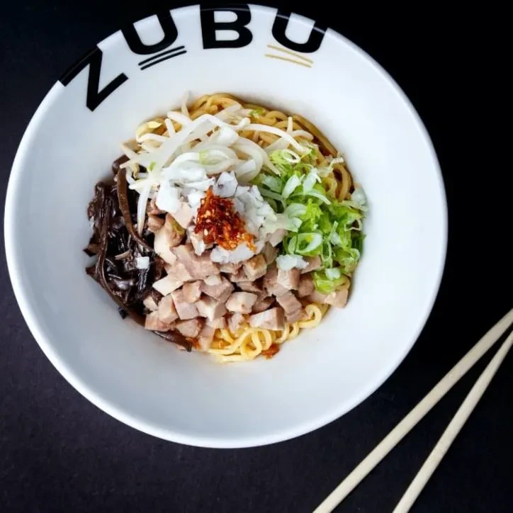 White ZUBU ramen bowl with noodles, meat, vegetables, and chopsticks.