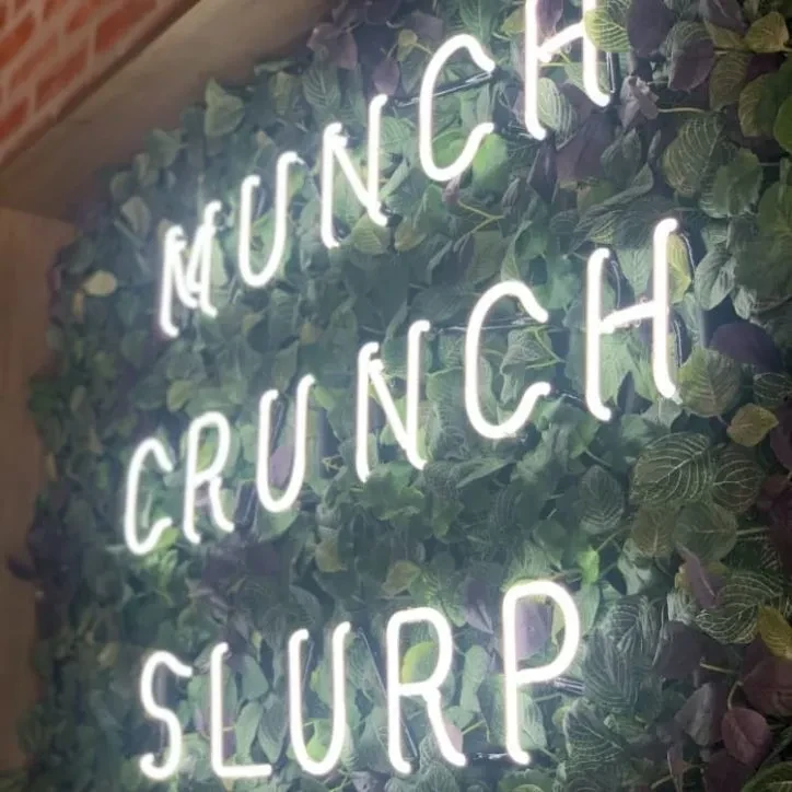 White neon sign "MUNCH CRUNCH SLURP" on artificial green foliage.