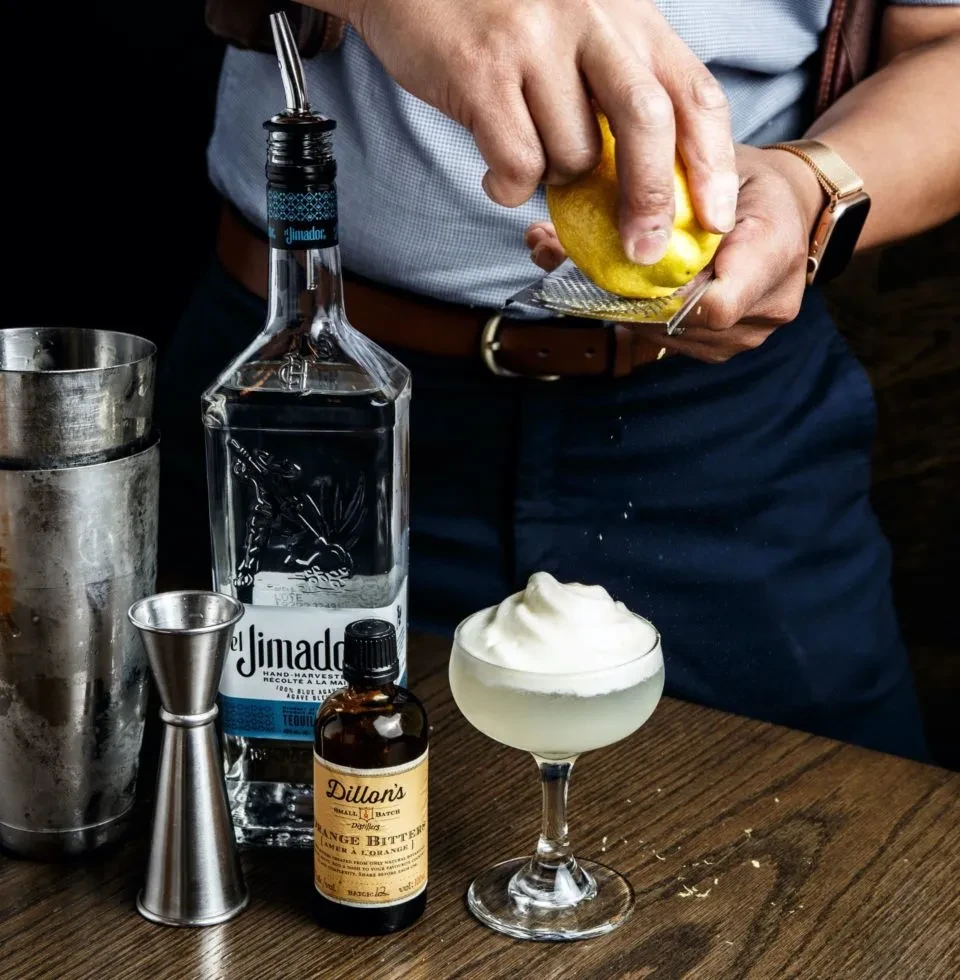 Bartender's hands zest lemon over foamy cocktail with tequila and bitters.